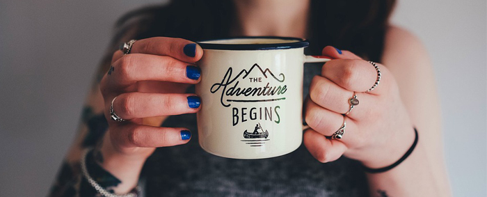 Women with tattoos holds a mug with the caption 'The Adventure Begins'