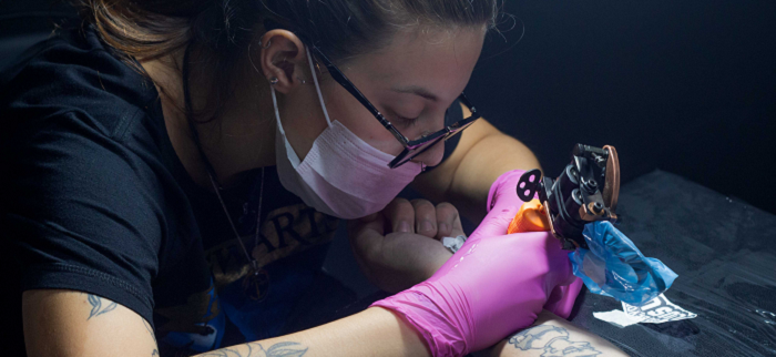 Female tattoo artists with glasses and face mask working.
